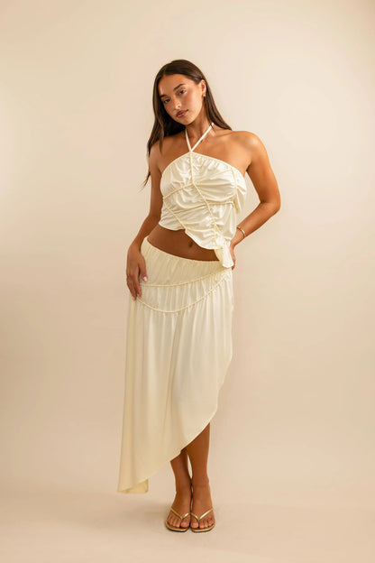 Koster Kulture Fleur Top and Skirt Set in cream featuring halter neckline crop top and asymmetrical midi skirt with sculpted piping detail and elegant silhouette